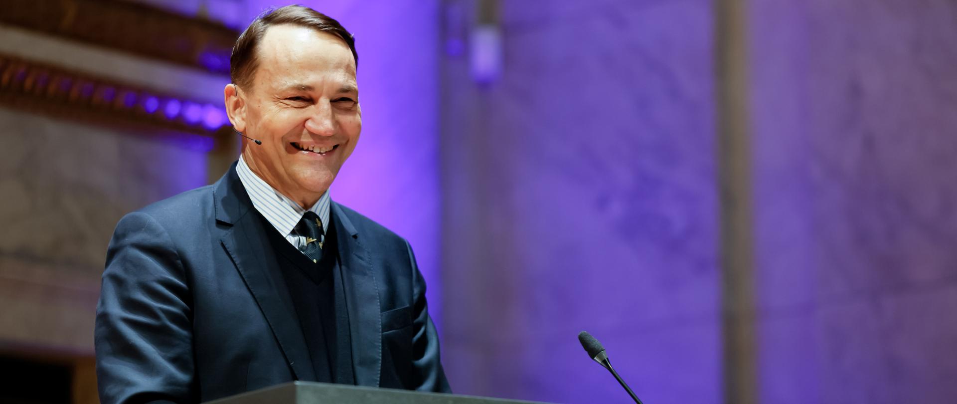 Mężczyzna w garniturze stoi przy mównicy.