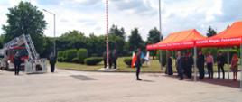 Fotografia przedstawia na pierwszym planie po prawej stronie funkcjonariuszy państwowej straży pożarnej w umundurowaniu wyjściowym stojących pod namiotem wraz z zaproszonymi gośćmi. Na środku w oddali widać poczet flagowy składający się z trzech strażaków stojących obok masztu z w zawieszoną flagą. W oddali po lewej stronie widać dwóch strażaków w umundurowaniu wyjściowym, a między nimi znajduje się wysunięty kosz samochodu pożarniczego specjalnego drabiny.