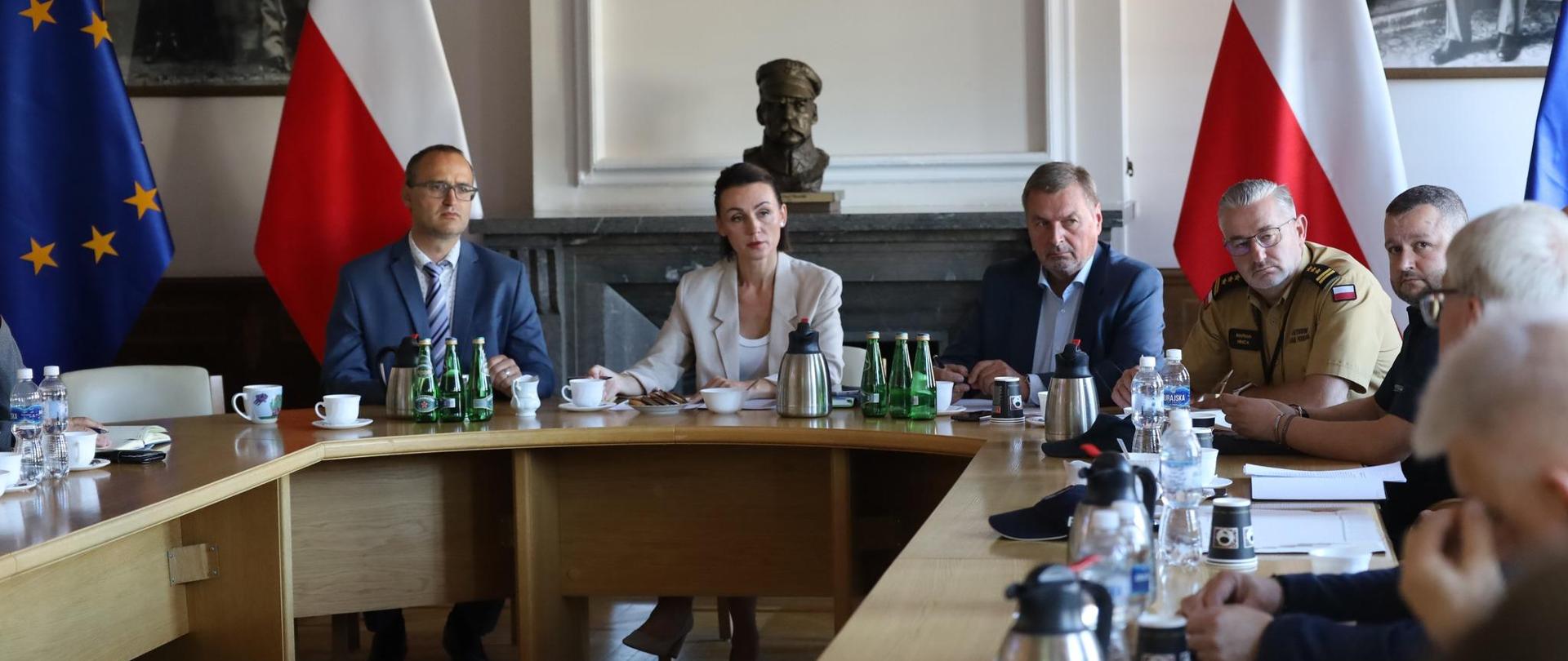 przy okrągłym stole siedzi kilka osób, w tle flagi polskie i unijne a także popiersie marszałka Piłsudskiego