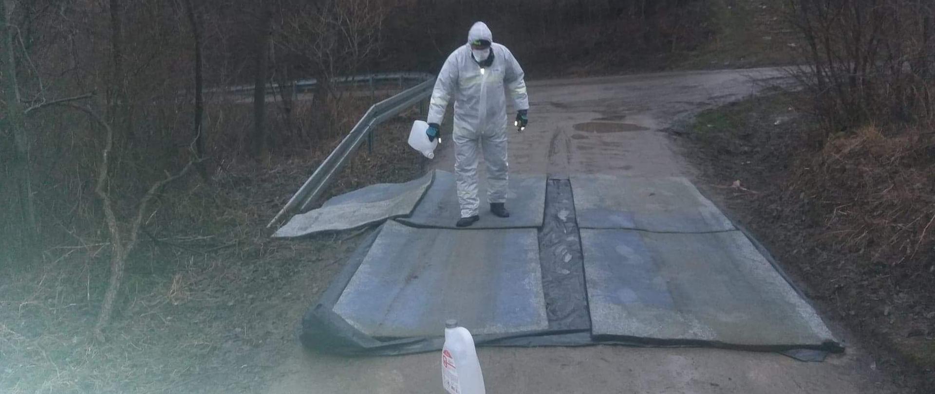 Osoba w specjalnym kombinezonie nasącza płynem maty dezynfekcyjne.