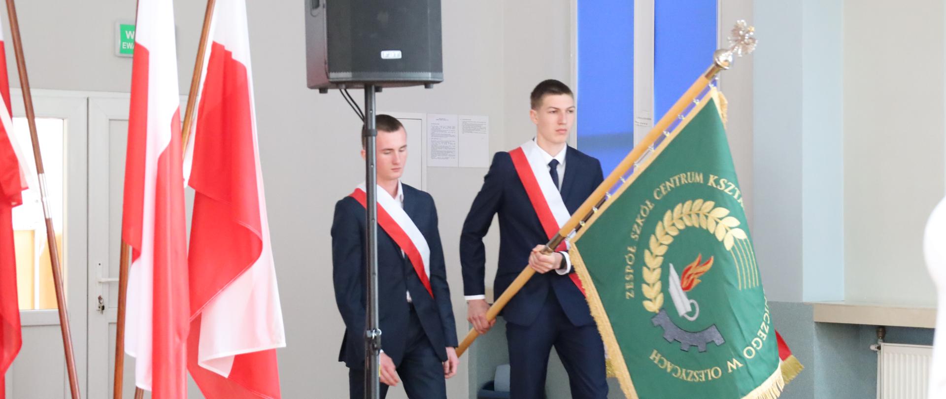 Trzech uczniów ubranych na galowo wprowadza sztandar szkoły.
