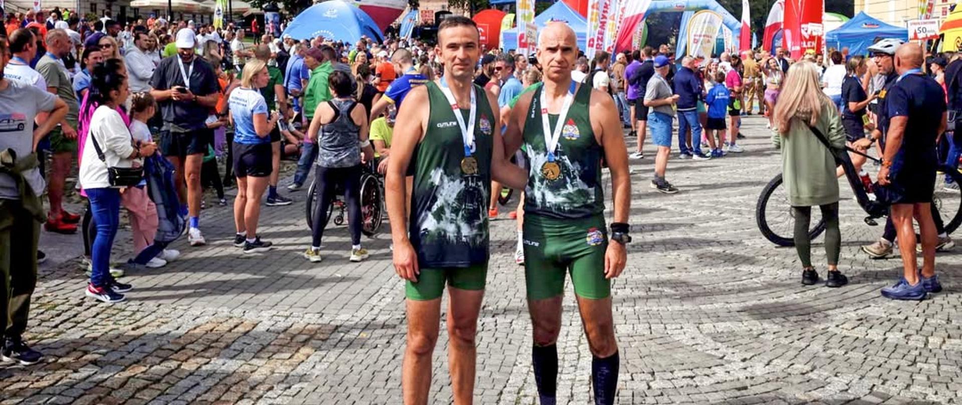 Zdjęcie przedstawia dwóch strażaków z KP PSP w Ustrzykach Dolnych w strojach sportowych przy starcie biegu. Wokół znajdują się postronni ludzie. 
