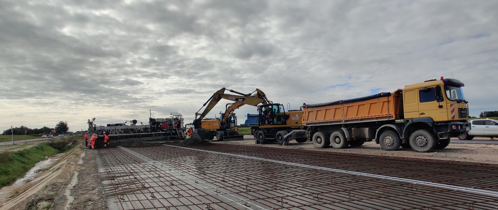 Na zdjęciu widoczny fragment zbrojenia ciągłego, po prawej stronie stoi pomarańczowa wywrotka. W części centralnej ustawiony zestaw maszyn do układania nawierzchni betonowej, po lewej stronie stara nitka drogi s7 po której poruszają się pojazdy osobowe.