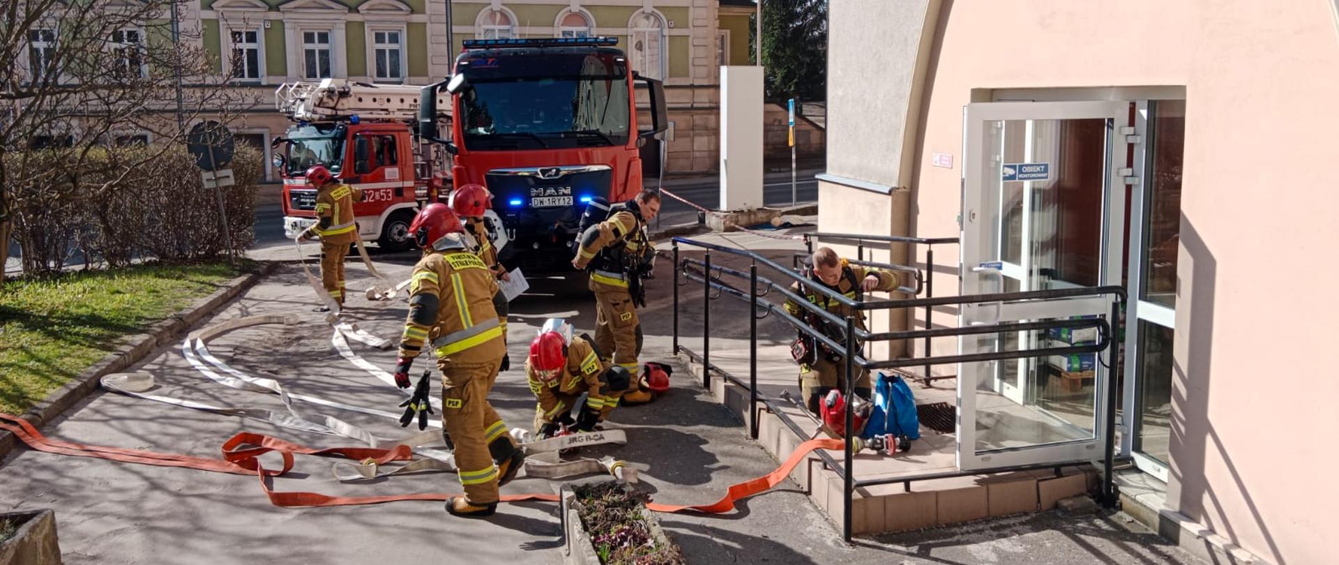Przed wejściem do budynku sześciu strażaków przygotowuje się do przeprowadzenia ćwiczeń. Za nimi stoją dwa pojazdy strażackie. W tle widok na dwukondygnacyjny budynek.