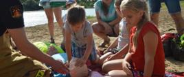 Piknik "Kręci mnie bezpieczeństwo nad wodą". Na kocu 3 manekiny do nauki zasad udzielania pierwszej pomocy, a przy nich klęczący strażak, który instruuje w jaki sposób prowadzić zewnętrzny masaż serca i sztuczne oddychanie. Wokół czworo dzieci i trzy osoby dorosłe. Dwie z nich stoją. Jedna z dziewczynek uciska ręką klatkę piersiową manekina, a drugą odchyla jego głowę. W tle akwen i drzewa.
