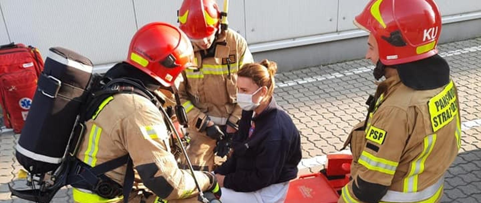 Trzech strażaków w ubraniach specjalnych rozmawia z kobietą siedzącą na noszach. Wspomniana grupa osób znajduje się na dworze. W tle widać ścianę budynku hali produkcyjnej.