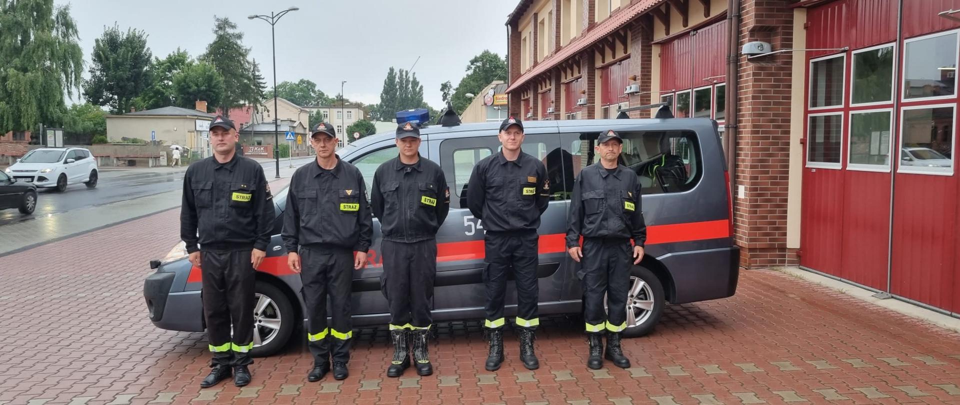 Zdjęcie przedstawia 5 strażaków, którzy uczestniczą w odprawie dotyczącej wyjazdu modułu GFFFV do walki z pożarami w Grecji. Na zdjęciu widzimy strażaków przed wyjazdem busem do punktu koncentracji sił i środków. Zdjęcie wykonane w porze dziennej przed placem Komendy.