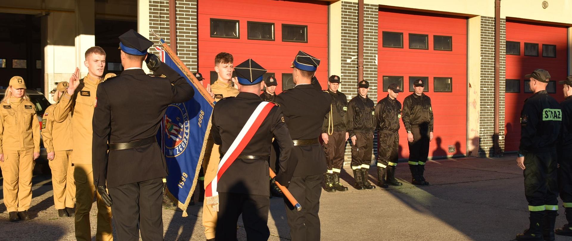Dzień. Przed bramami garażowymi w kolorze czerwonym stoi kilkunastu funkcjonariusz PSP ustawionych w dwuszeregu. Obok nich przy poczcie sztandarowym reprezentującym sztandar Komendy Powiatowej PSP w Zgorzelcu stoi dwóch funkcjonariuszy. Obydwaj mają podniesioną prawą rękę w geście ślubowania