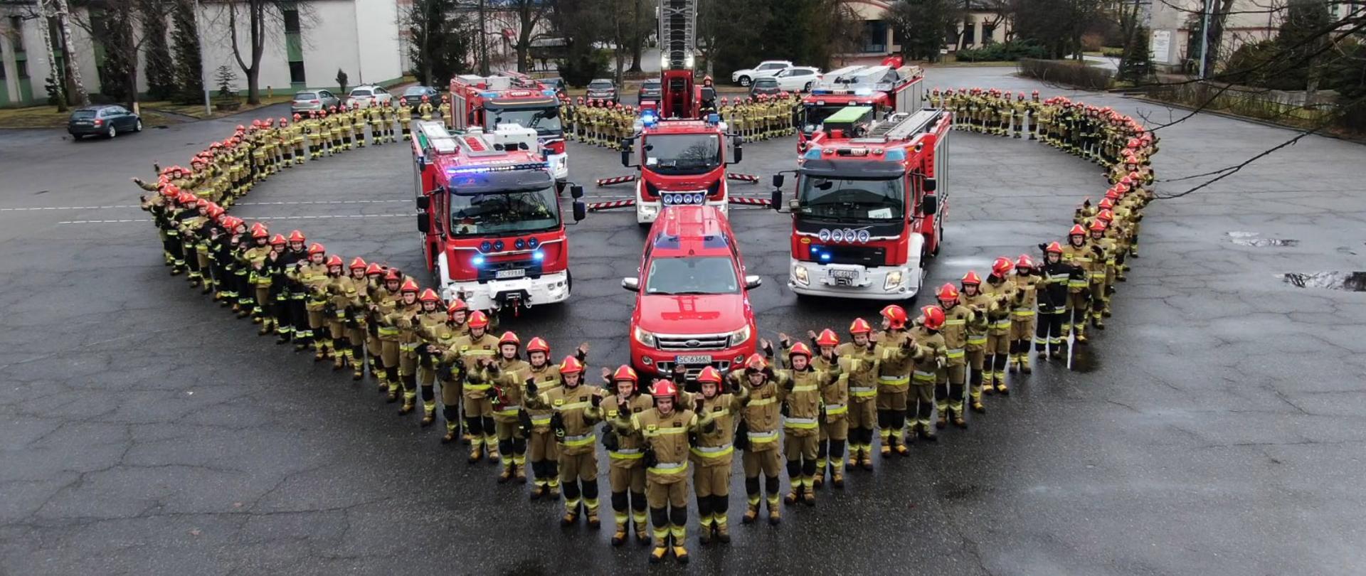 Plac wewnętrzny Szkoły, na którym zostało ustawionych 6 samochodów strażackich. Samochody otaczają dwie kompanie kadetów, tworząc figurę w kształcie serca