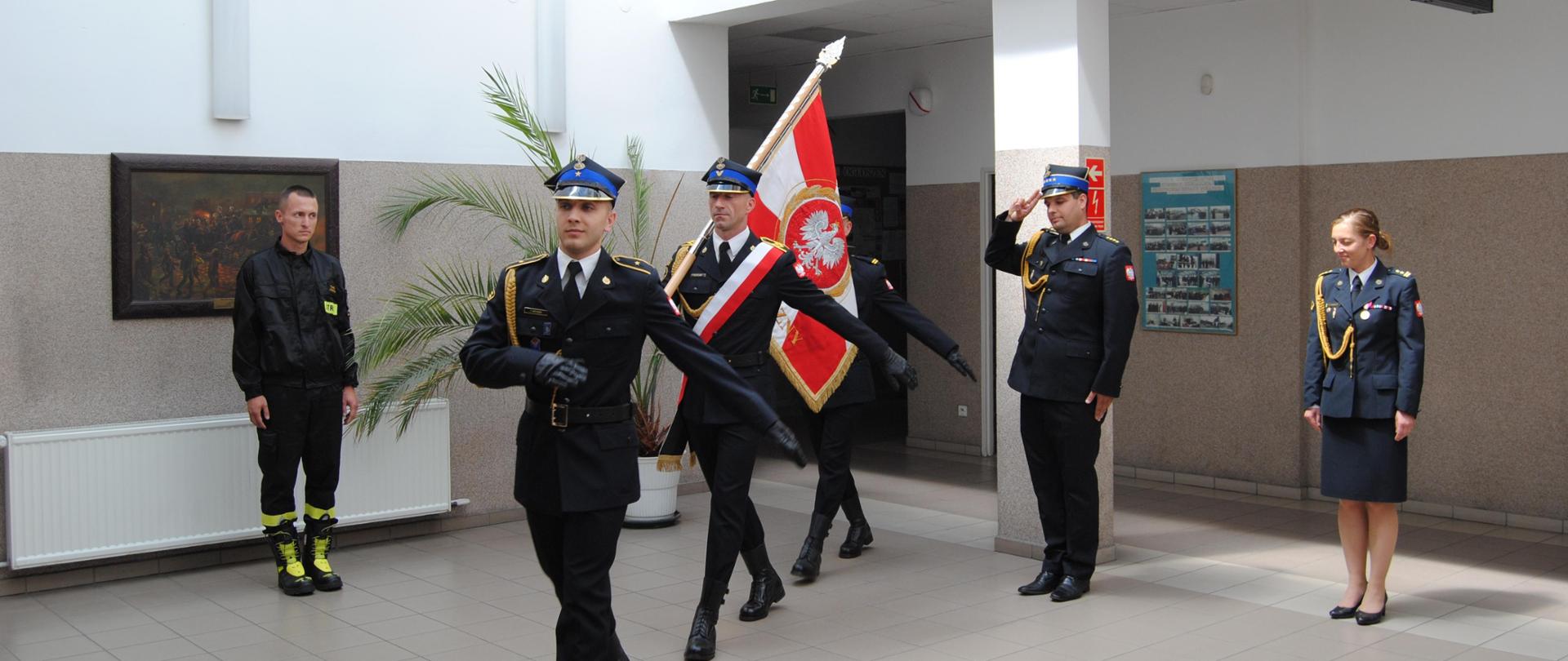 W pomieszczeniu maszeruje jeden za drugim trzech strażaków w mundurach reprezentacyjnych, środkowy niesie na ramieniu sztandar. Po lewej stoi na baczność w ubraniu koszarowym. Po prawej salutuje dowódca JRG, dalej Pani Komendant.