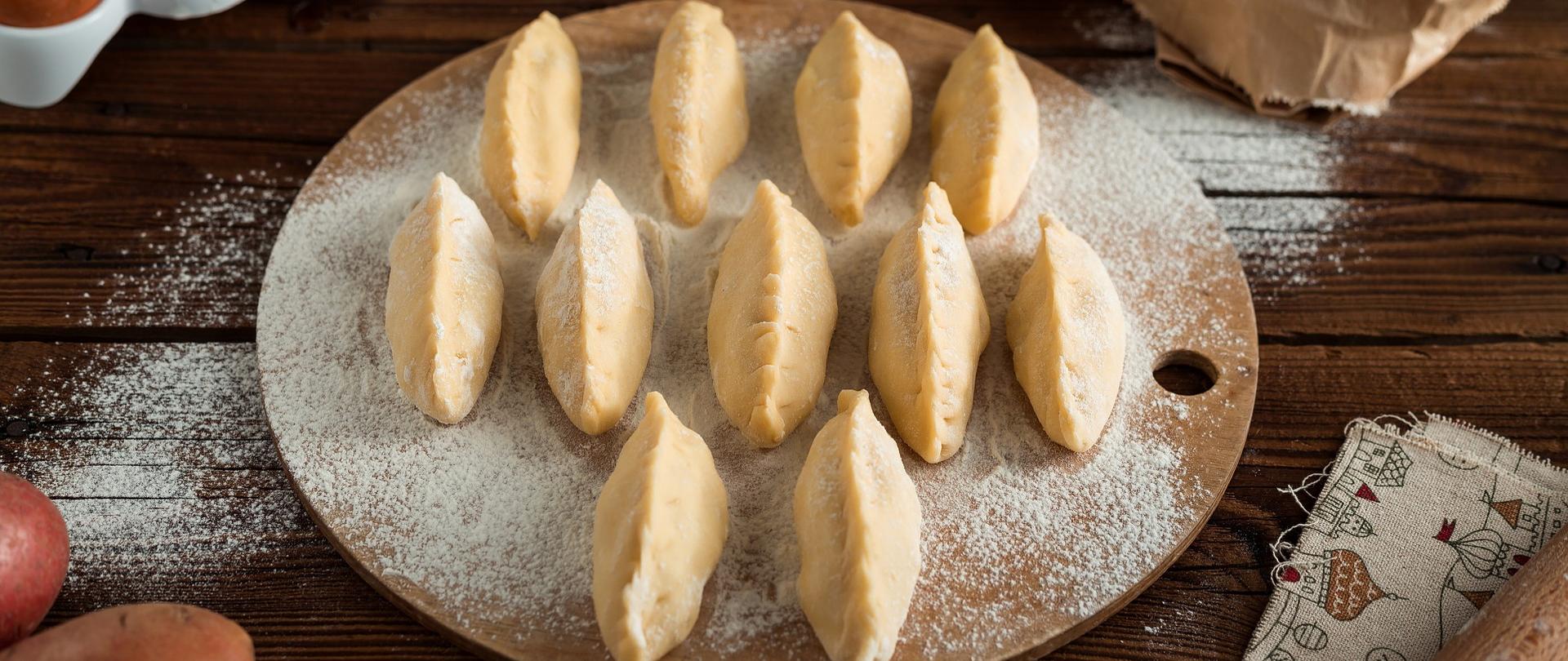 Na środku zdjęcia na drewnianej okrągłej tacy są pierogi obsypane mąką. W lewym dolnym rogu są dwa ziemniaki. W lewym górnym rogu są jajka. W prawy górnym rogu jest mąką.