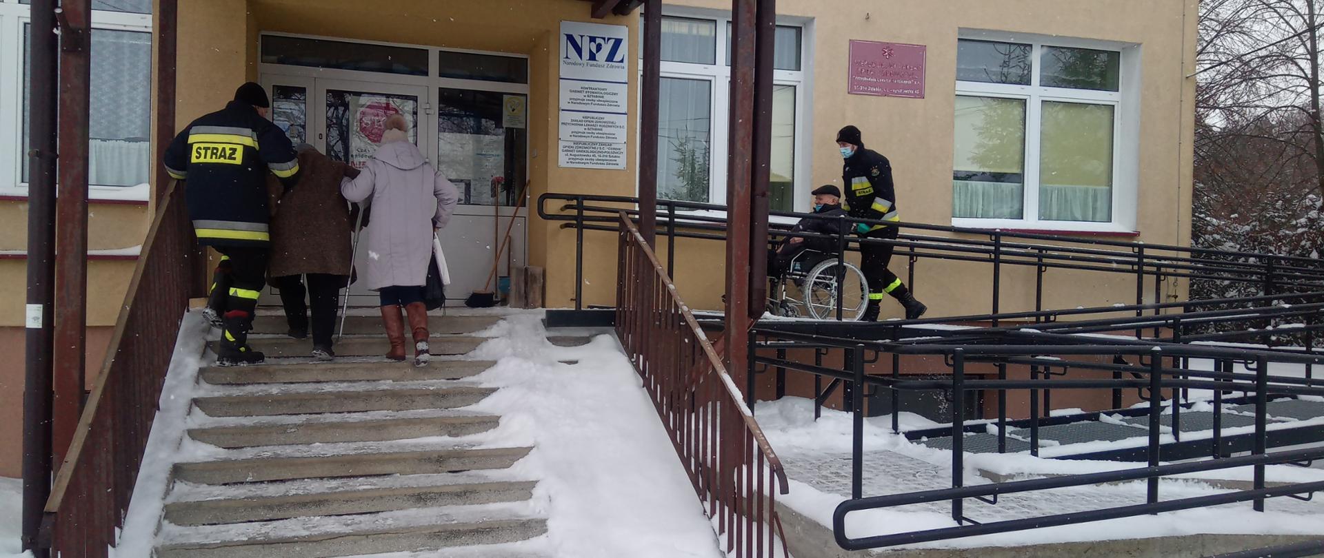Na zdjęciu widać jak jeden strażak przemieszcza na wózku inwalidzkim jedną osobę do punktu szczepień w msc. Sztabin, drugi strażak wprowadza po schodach (z osobą postronną) drugą osobę.