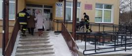 Na zdjęciu widać jak jeden strażak przemieszcza na wózku inwalidzkim jedną osobę do punktu szczepień w msc. Sztabin, drugi strażak wprowadza po schodach (z osobą postronną) drugą osobę.