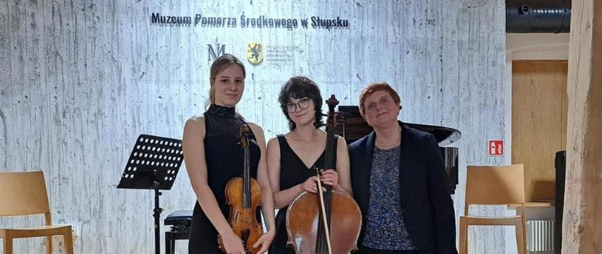 Zdjęcie kolorowe. Po koncercie, przed sceną stoją razem skrzypaczka, wiolonczelistka i ich akompaniatorka. W tle fortepian, krzesła i pulpity.