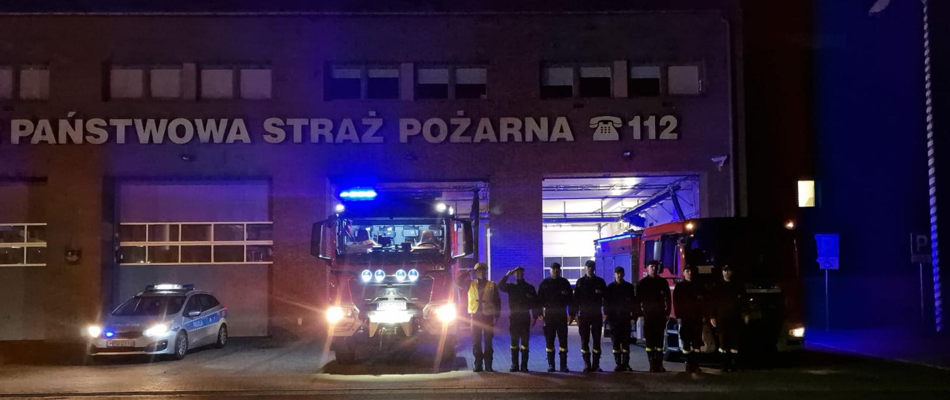 Na tle komendy straży pożarnej samochody pożarnicze i radiowóz policji przed samochodami stoją funkcjonariusze oddając honory