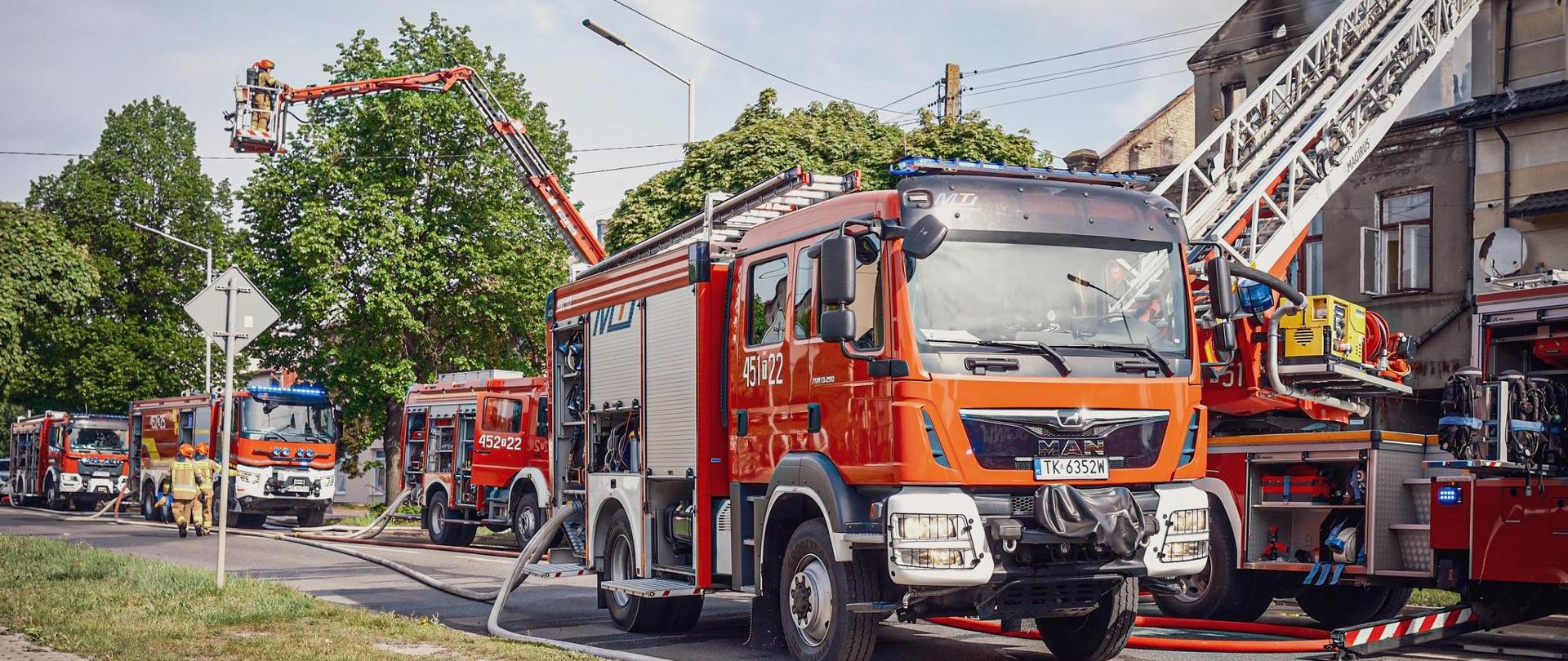 Na zdjęciu widoczne są działania strażaków, widać na nim samochody gaśnicze oraz strażaków na ziemi a także na drabinie mechanicznej widać również wijące sie po ziemi węże dostarczające wodę do gaszenia.