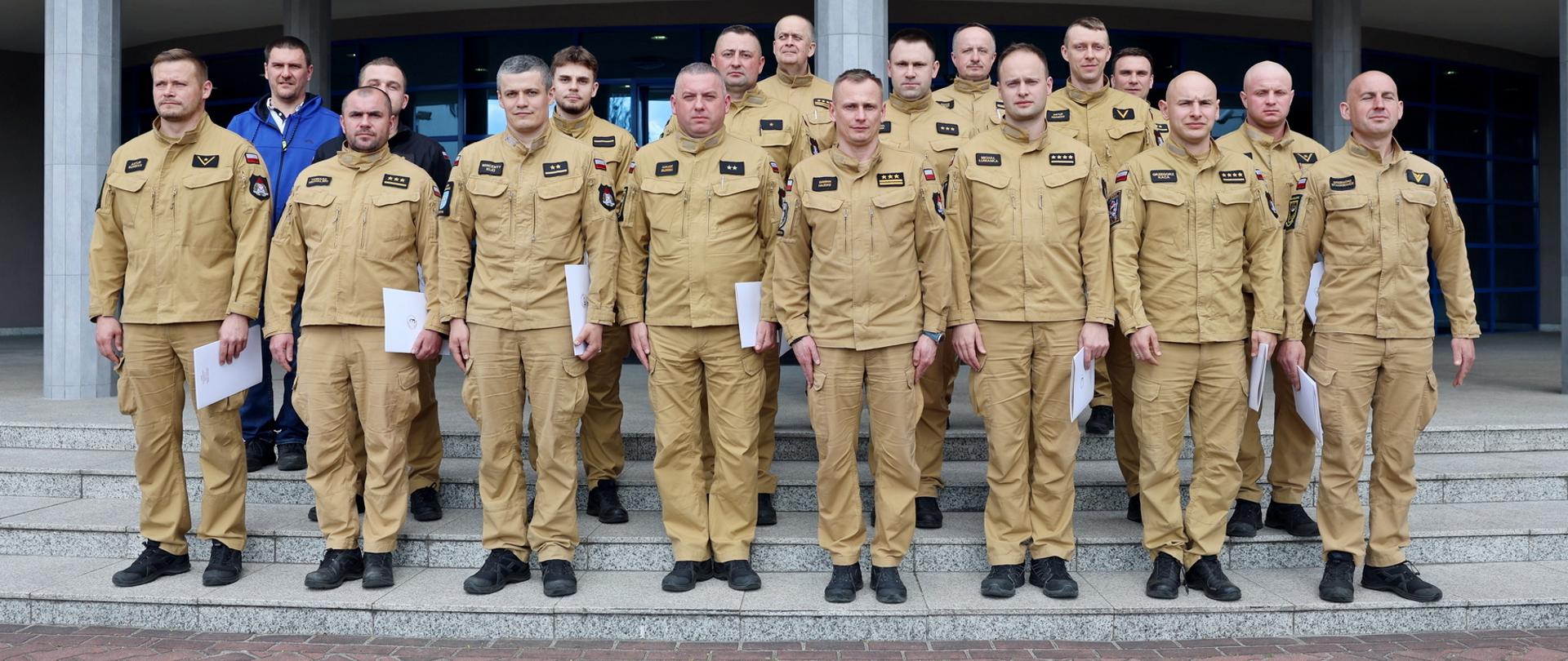 Pamiątkowe zdjęcie grupowe przed budynkiem Centralnej Szkoły PSP. Kilkunastu strażaków w jednakowych piaskowych mundurach stoi na schodach przed wejściem do budynku, trzymając dokumenty potwierdzające ukończenie szkolenia. Wszyscy ustawieni są w kilku rzędach i patrzą w stronę aparatu