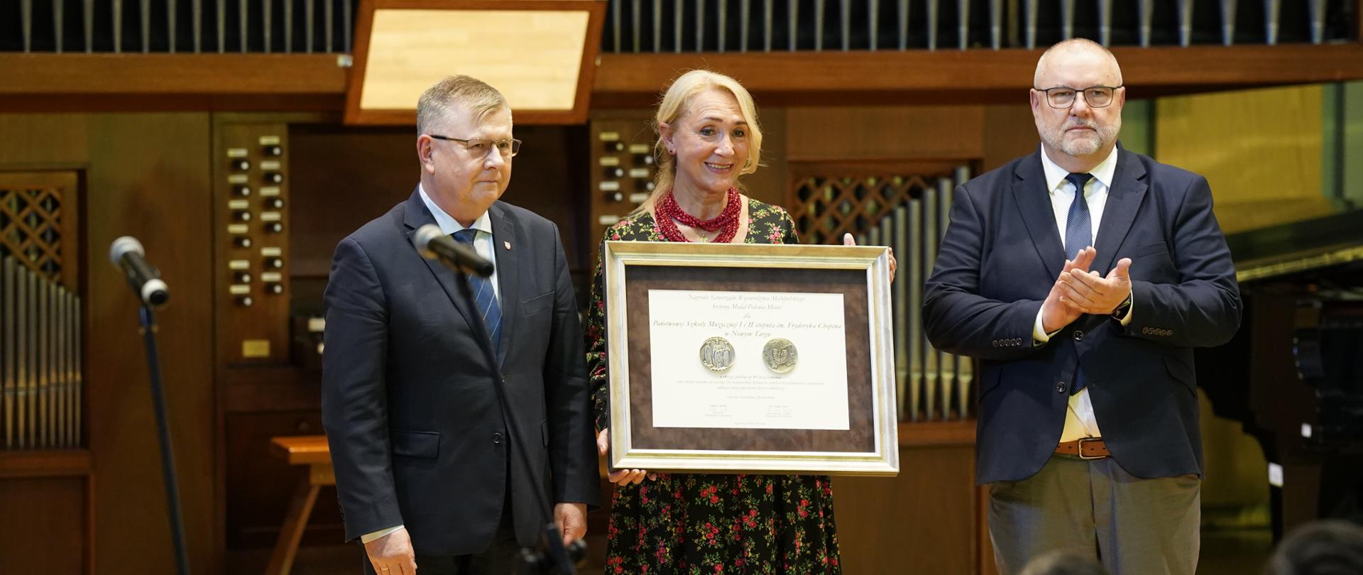 Zdjęcie przedstawia oficjalną uroczystość wręczenia nagrody lub wyróżnienia, która odbywa się na scenie, najprawdopodobniej w sali koncertowej lub filharmonii (w tle widoczne są duże organy piszczałkowe oraz fragment fortepianu). Na pierwszym planie stoją trzy osoby: Po lewej stronie: Mężczyzna w ciemnym garniturze, koszuli i krawacie, stojący tyłem do mikrofonu. W centrum: Uśmiechnięta kobieta w kwiecistej sukience i czerwonych koralach, która trzyma w dłoniach dużą, oprawioną w ramy antyramę. Wewnątrz widać dyplom oraz dwa pamiątkowe medale. Po prawej stronie: Mężczyzna w okularach, granatowej marynarce i szarych spodniach, który klaszcze w dłonie. Przed sceną widoczne są sylwetki publiczności oraz fotografów dokumentujących to wydarzenie. Całość ma elegancki, podniosły charakter.