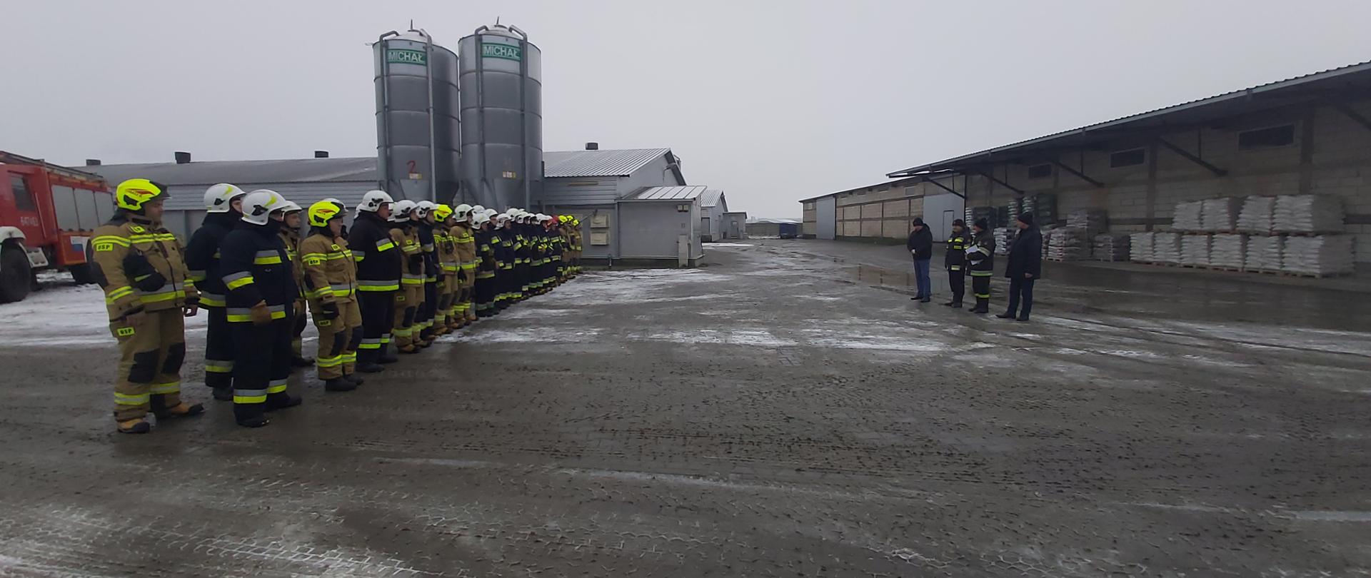 Gminne Ćwiczenia Ochotniczych Straży Pożarnych w miejscowości Stasin - za zdjeciu podczas zakończenia ćwiczeń strażacy w ubraniu specjalnym stoją w dwuszeregu na zbiórce na kostce, na niej leży śnieg, w tle budynki magazynowe