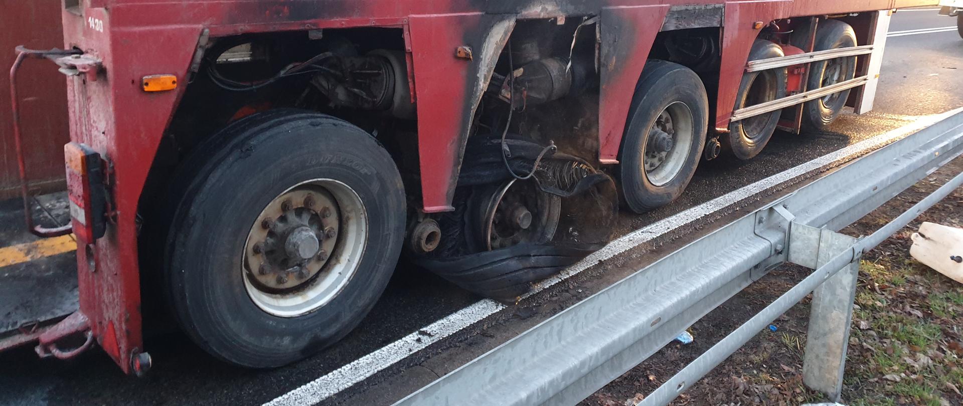 Na zdjęciu znajduje się załadowana naczepa samochodu ciężarowego do przewozu ładunków specjalnych, która stoi na drodze. Pożarem objęte zostało podwozie naczepy wraz z ogumieniem.