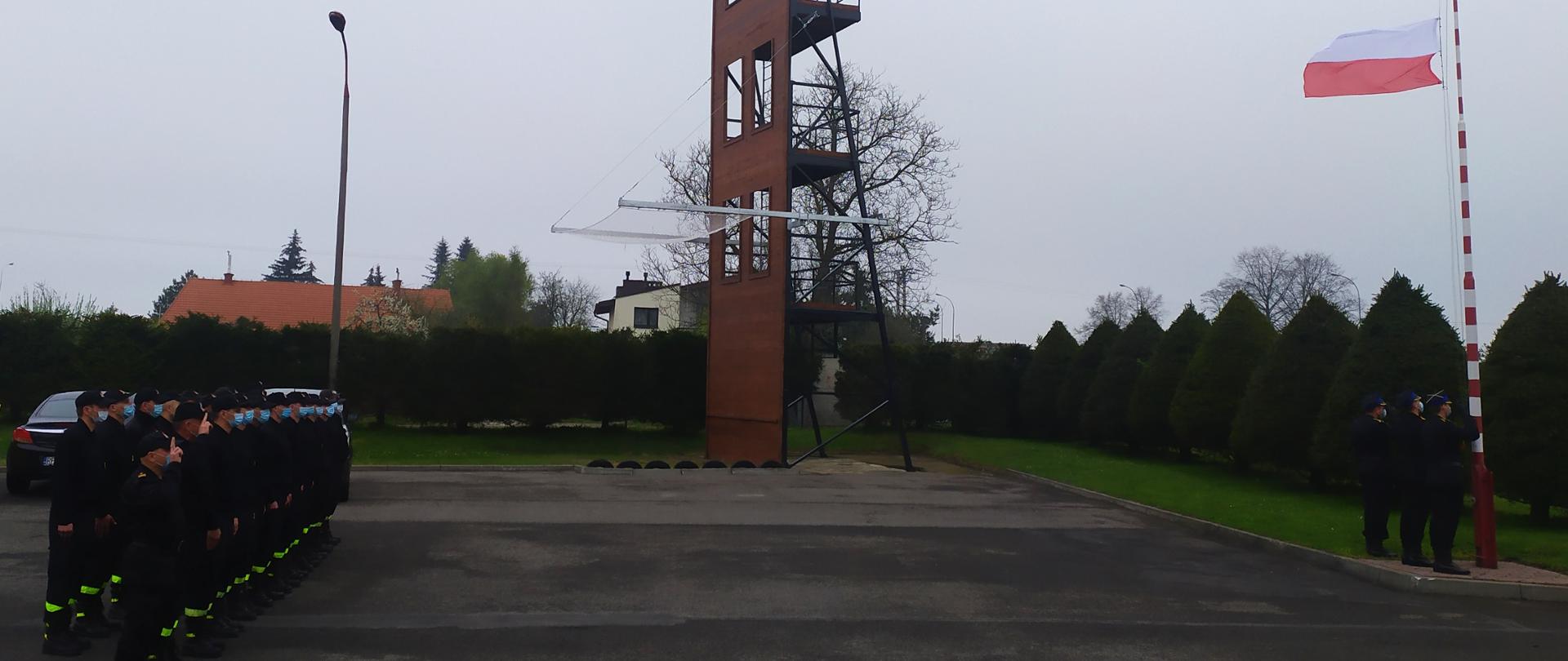 Na zdjęciu widać po lewej stronie pododdział funkcjonariuszy Komendy Powiatowej Państwowej straży Pożarnej w Jarosławiu, a po prawej podniesioną na maszcie flagę Polski i stojąc pod masztem poczet flagowy. Strażacy w pododdziale ubrani w umundurowanie koszarowe, a poczet flagowy w mundur wyjściowy. Wszyscy strażacy mają na twarzy maseczki.