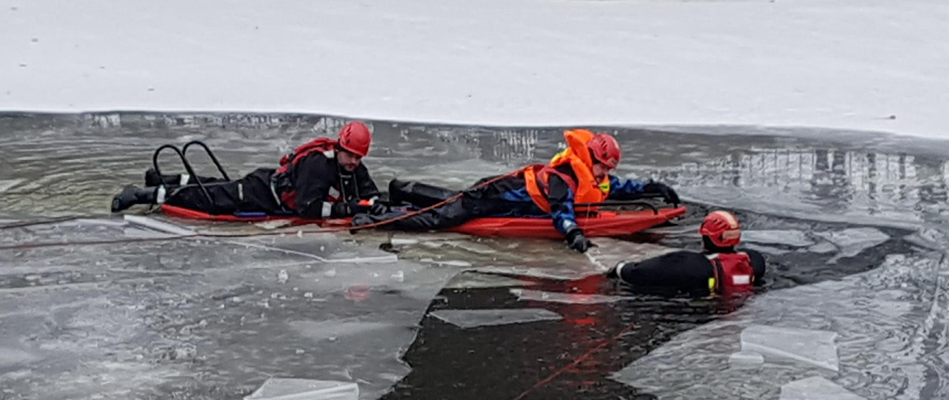 Dwójka strażaków na saniach lodowych przemieszczających się po zamarzniętym stawie w kierunku trzeciego strażaka zanurzonego w wodzie.