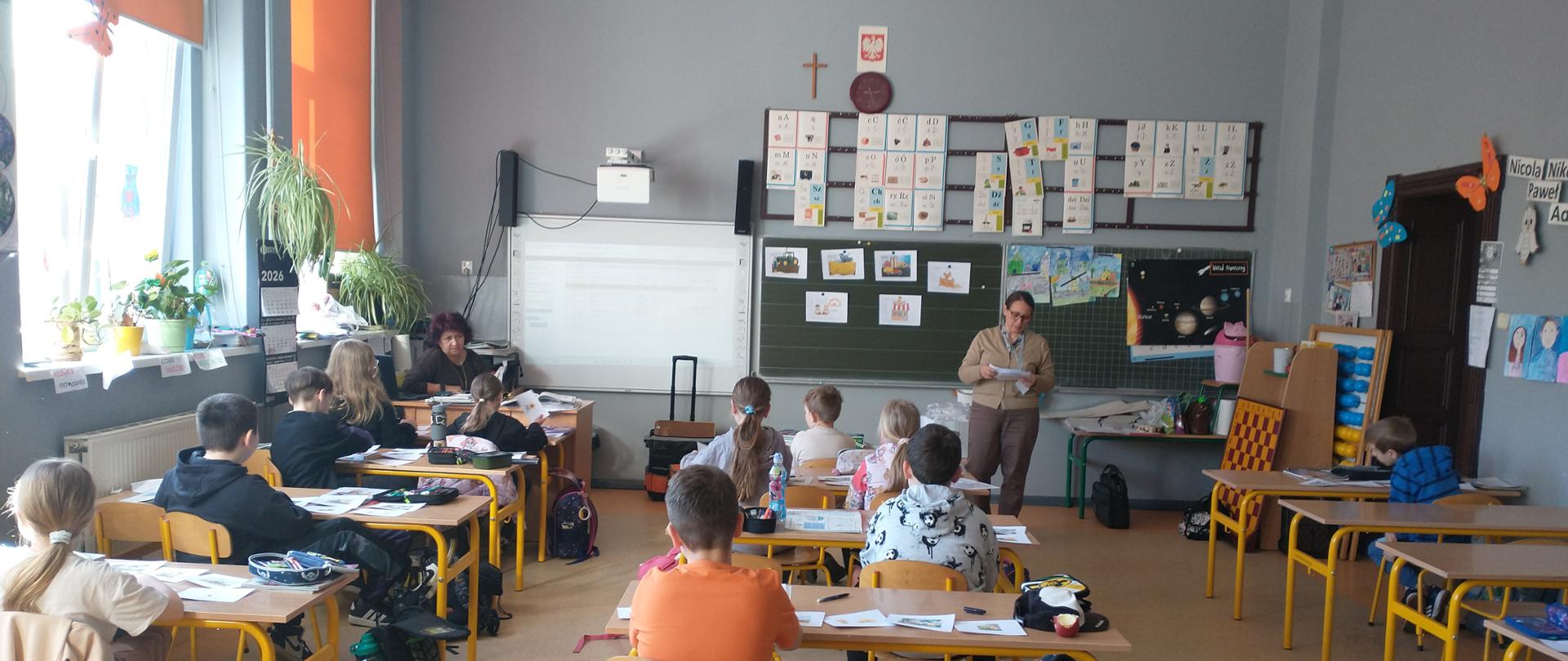 W klasie szkolnej dzieci siedzą przy pojedynczych ławkach i pracują na kartkach. Inspektor stoi z przodu przy tablicy i czyta z kartki. Na ścianie widać materiały edukacyjne, w tym plansze z literami i ilustracjami.