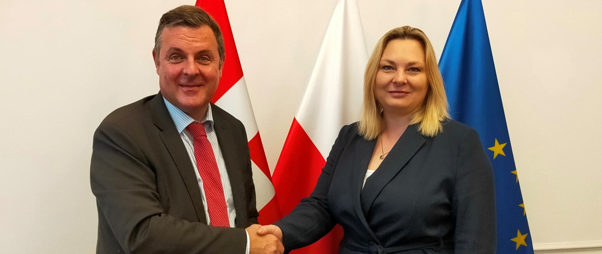 Wiceminister funduszy i polityki regionalnej Ewelina Owczarska i Patric Franzen – Zastępca sekretarza stanu ds. europejskich, Federalny Departament Spraw Zagranicznych Szwajcarii, pozują do zdjęcia na tle flag