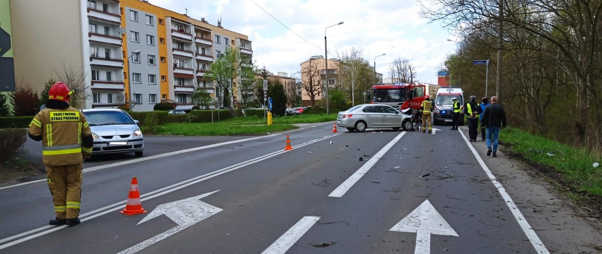 Na zdjęciu widoczne są dwa samochody osobowe oraz samochód ratowniczo-gaśniczy i karetka, strażacy oraz funkcjonariusze Policji prowadzący swoje działania. Dwie osoby postronne . Zdjęcie wykonane na tle osiedla.