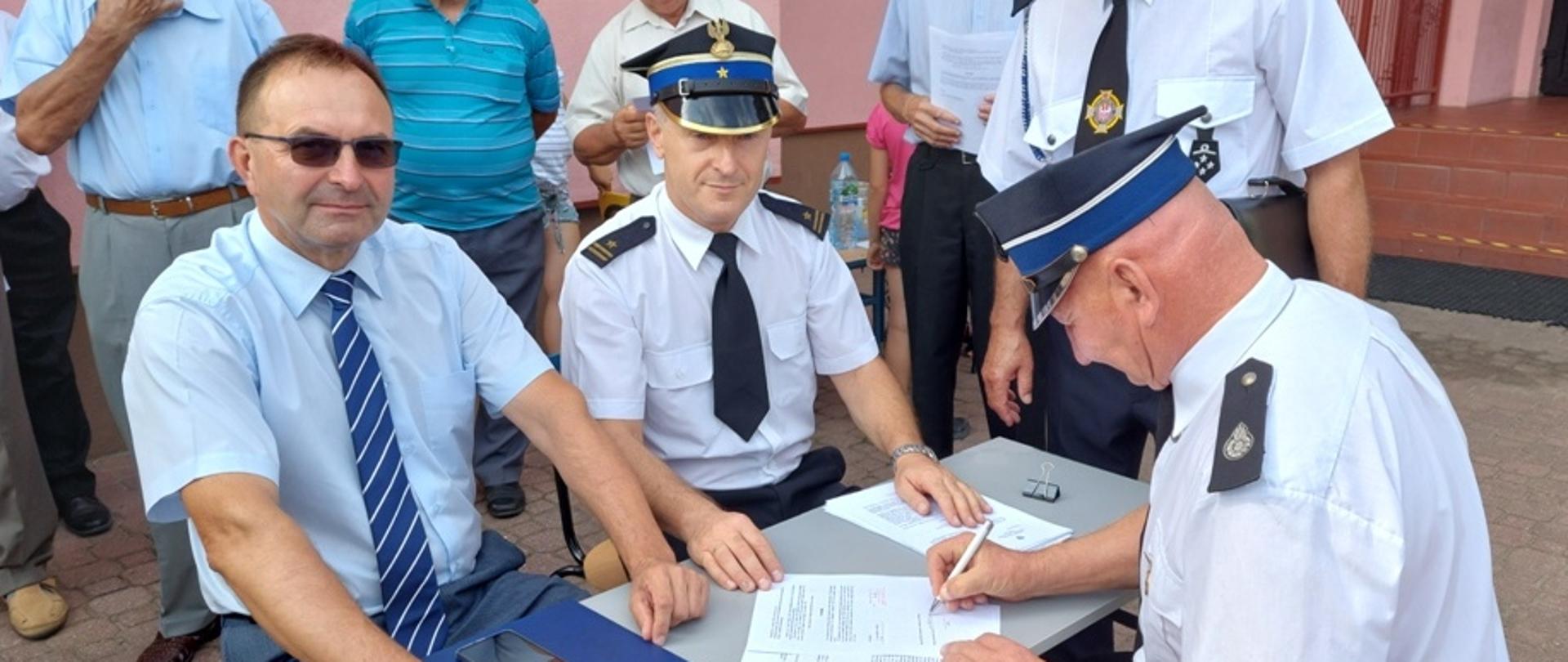 Kolorowe zdjęcie wykonane na zewnątrz przedstawia trzy osoby siedzące przy stoliku. Dwie osoby w mundurach Państwowej Straży Pożarnej i Ochotniczej Straży Pożarnej oraz jedna osoba cywilna. Jedna z nich podpisuje dokumenty.