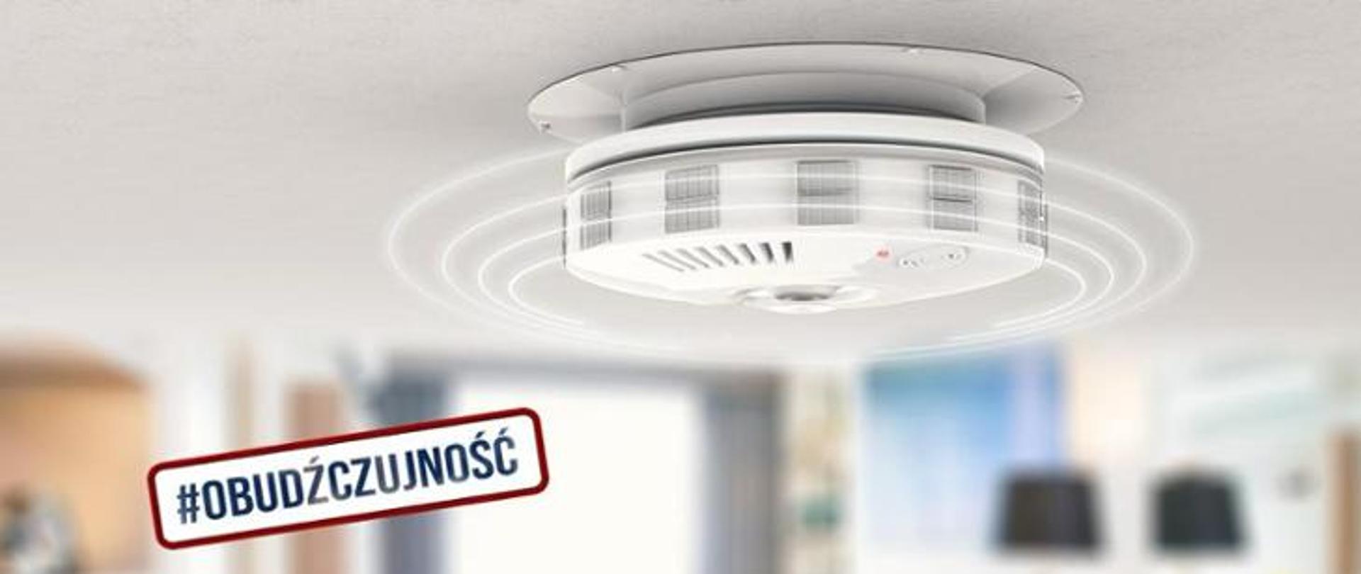 Grafika przedstawiająca opis: „Obudżczujność” oraz czujkę tlenku węgla zamontowaną na suficie pomieszczenia. 