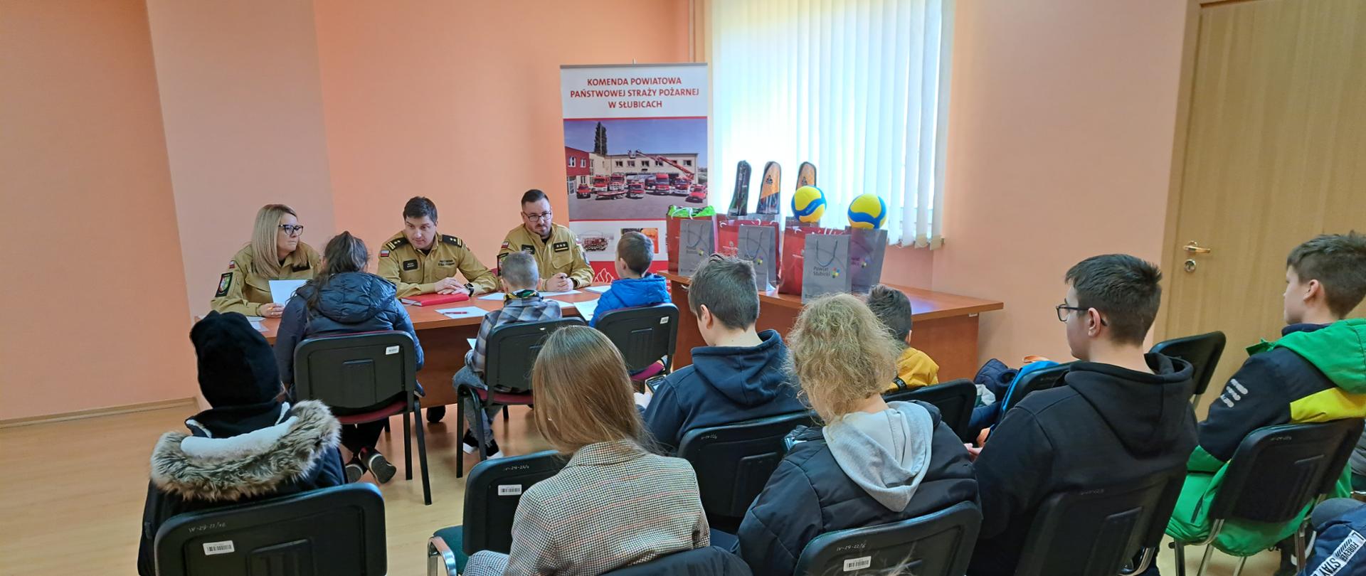 Wnętrze świetlicy KP PSP w Słubicach. Przy stole siedzą umundurowana trójka strażaków PSP stanowiących komisję turnieju. Przed nimi trójka dzieci odpowiadających na pytania. Na sali na krzesłach inne osoby obserwujące turniej.