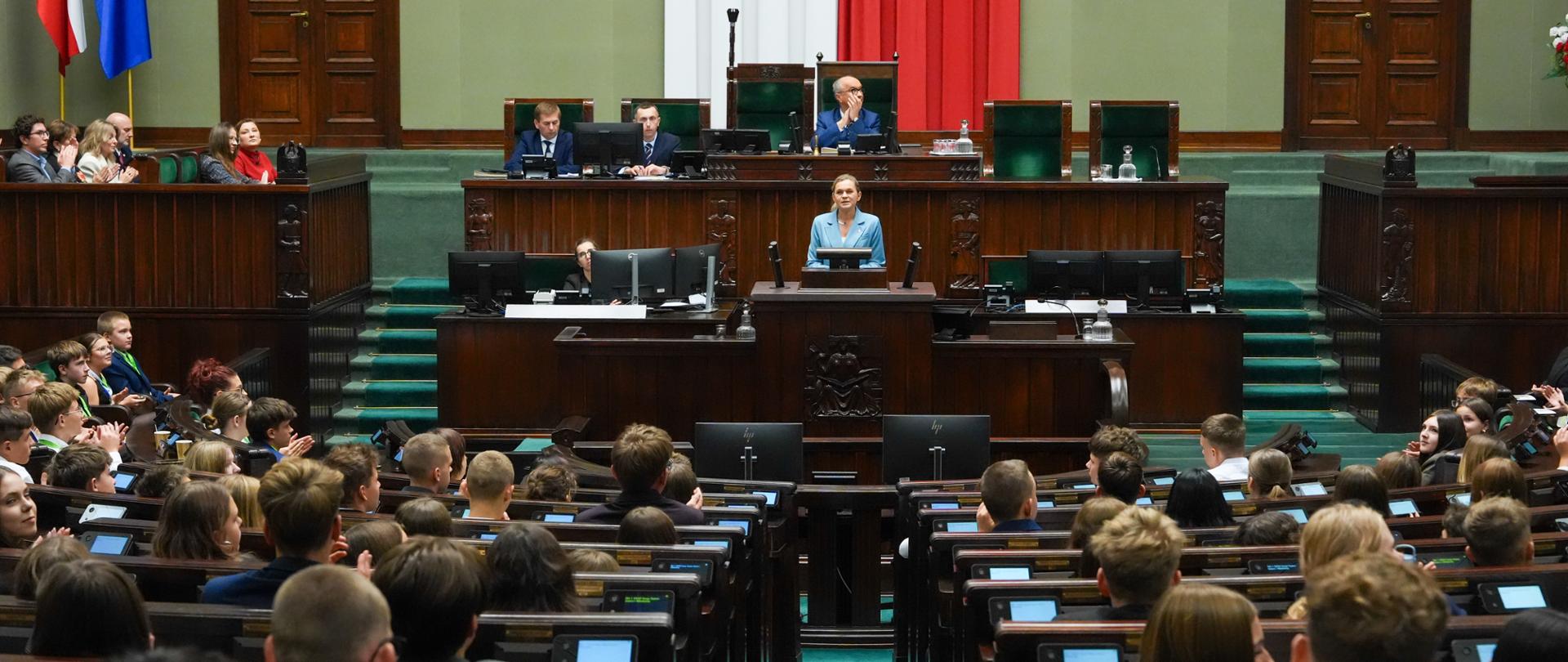 Szerokie ujęcie sali posiedzeń Sejmu z perspektywy ław poselskich w trakcie przemówienia minister Barbary Nowackiej stojącej przy ambonce.