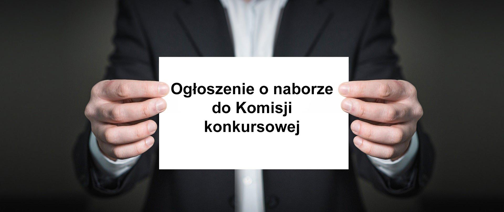Na zdjęciu męskie dłonie wyciągnięte w kierunku kadru z białą kartką, na której napisano "ogłoszenie o naborze do komisji konkursowej". W dalszym planie pozostała część tułowia. 