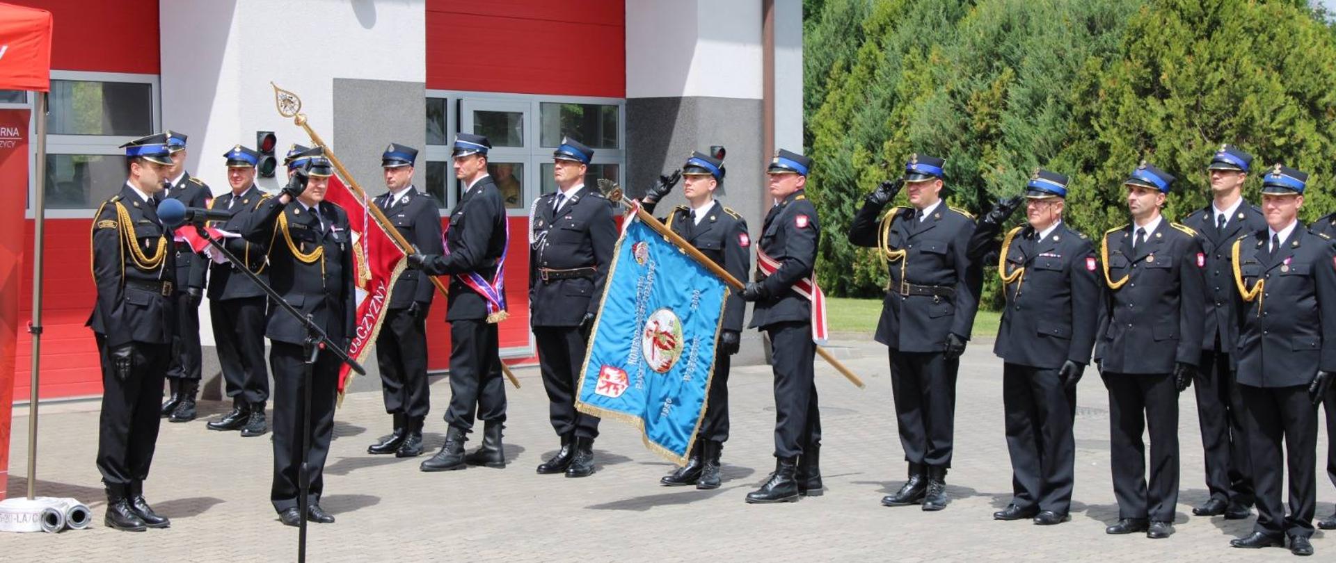 Na zdjęciu widać strażaków w galowych mundurach podczas uroczystego apelu przed budynkiem komendy. Część z nich salutuje, a dwóch trzyma sztandary jednostki.