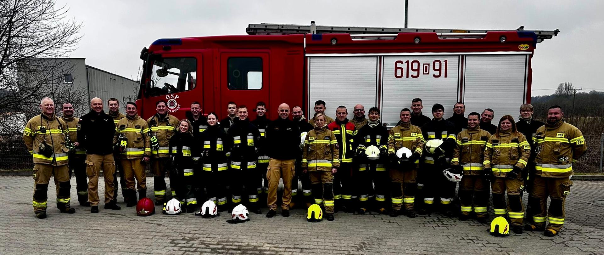duża grupa strażaków stoi w ubraniach specjalnych na tle samochodu pożarniczego