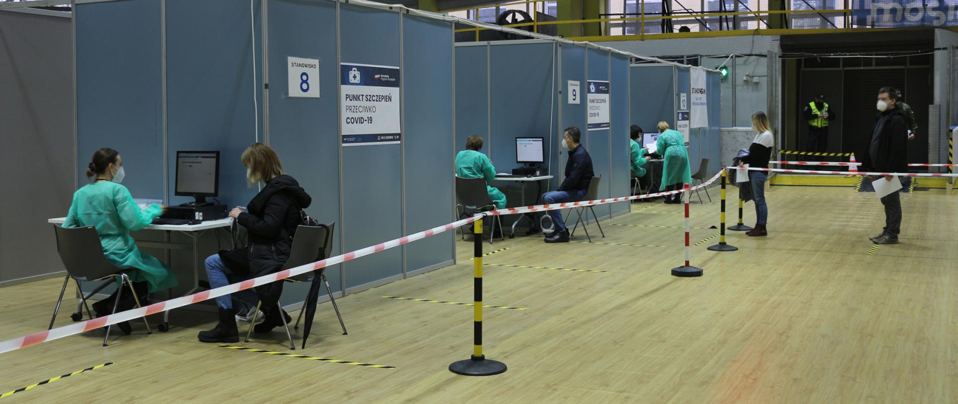 Na zdjęciu Punkt Szczepień Powszechnych w Opolu. Fotografia z wnętrza punktu. Na środku kadru widać boksy - a w nich osoby, zasiadające przed komputerem. To rejestratorzy szczepień. Obok nich na krzesłach siedzą pacjenci, przybyli na szczepienia. Przed boksami widać pachołki i biało-czerwoną wstęgę, oddzielającą sektory. Po prawej stronie kadru widać kolejkę pacjentów, oczekujących na rejestrację. Na górze kadru w jego centralnej części widać tablicę na której wyświetla się godzina 8:33. 