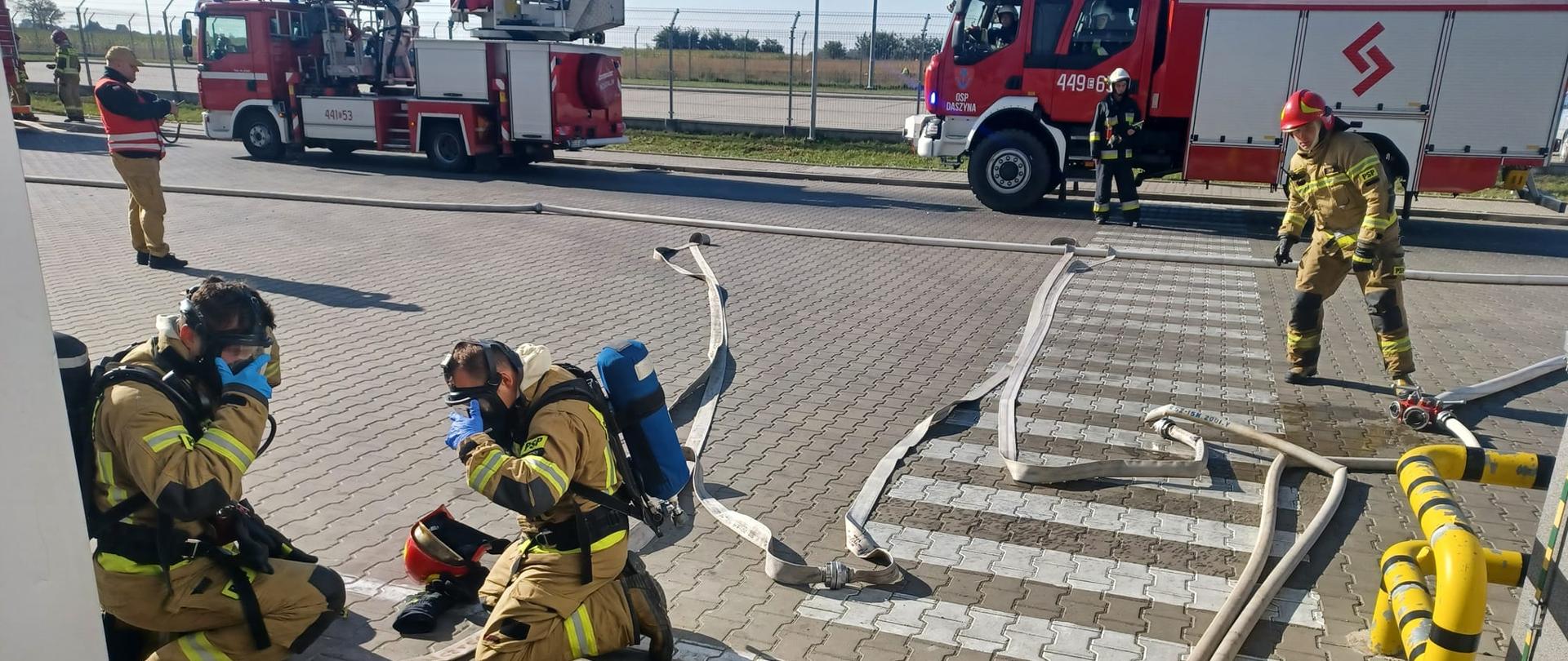 Na zdjęciu widać strażaków, którzy rozwijają węże strażackie oraz ubierają się w maski do aparatów ochrony dróg oddechowych. Na ziemi leżą szare węże strażackie. W tle stoją dwa samochody strażackie. Jest widno.