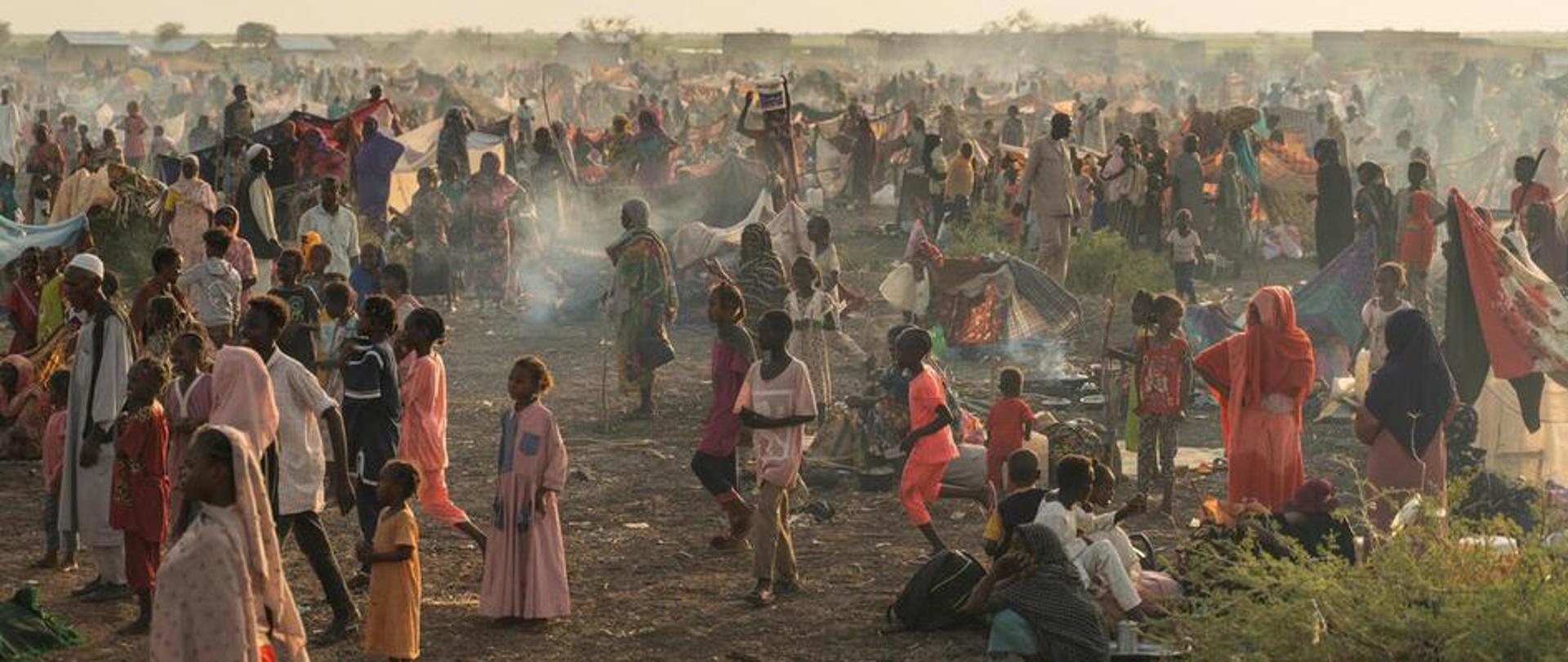 Tłum ludzi w różnym wieku zebrany na zakurzonym polu z kilkoma niskimi domami na horyzoncie.