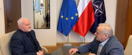 2 mężczyzn siedzi na przeciwko siebie, siedzą przy stole, rozmawiają. W tle widać 3 flagi - polską, UE i NATO