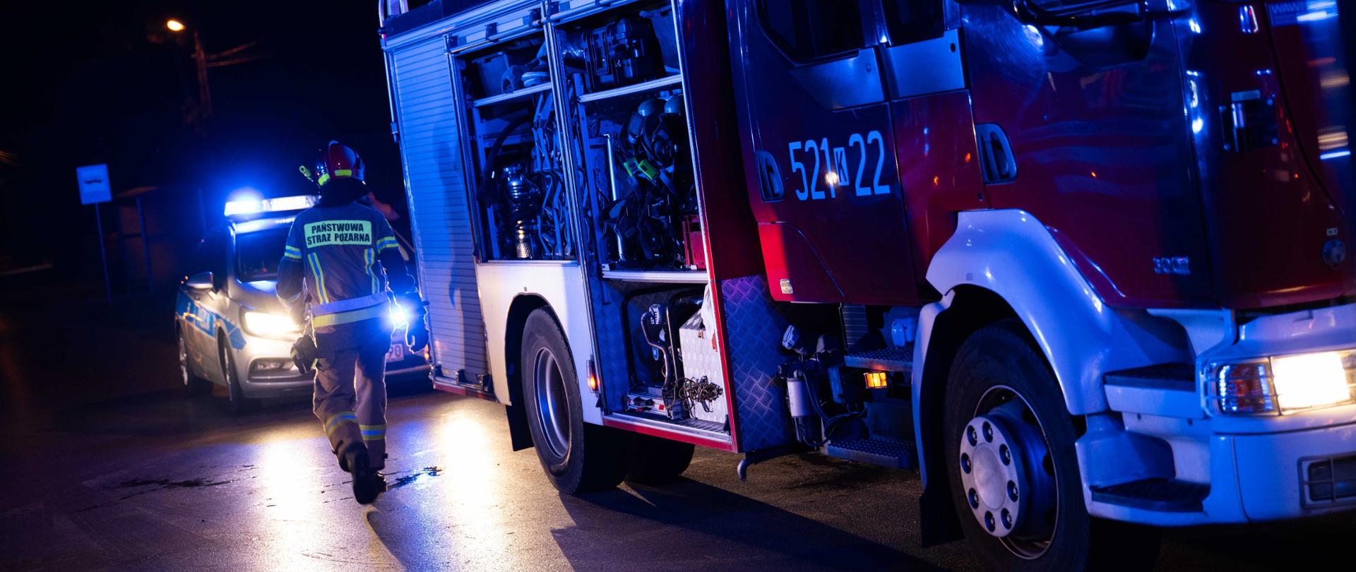Strażak stoi tyłem a w tle samochód pożarniczy oraz rozbite auto