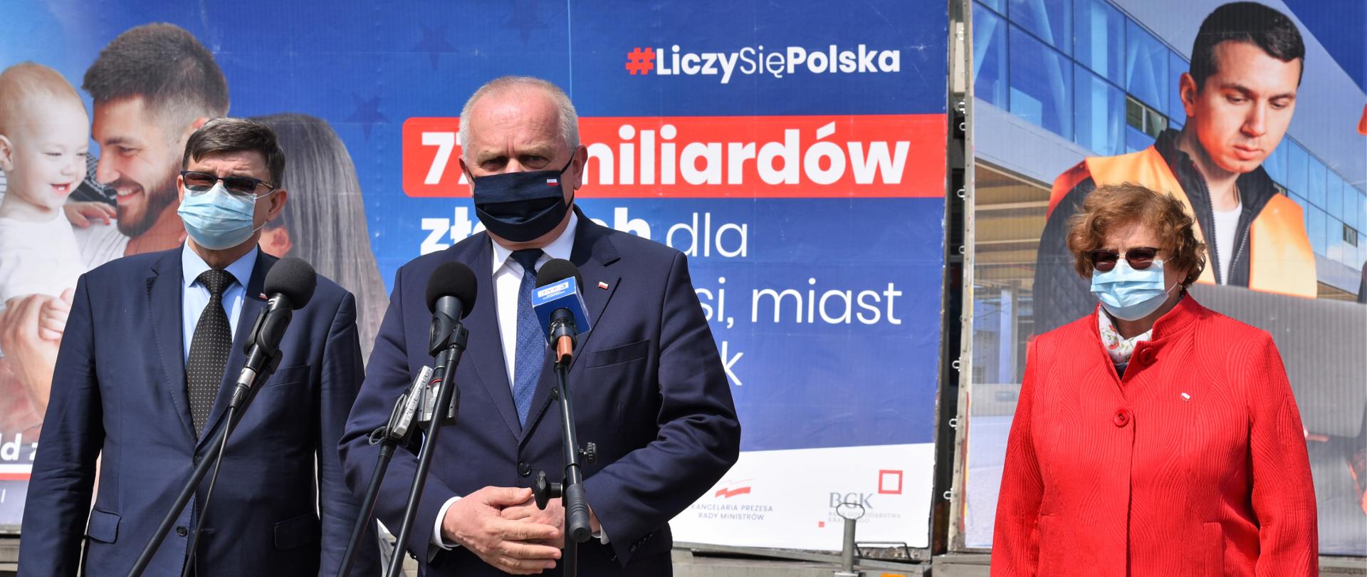 Wojewoda przemawia na tle plansz promocyjnych Liczy się Polska. Przed nim ustawione mikrofony. Za nim, po obu stronach stoją Poseł Elżbieta Płonka i Poseł Jerzy Materna 