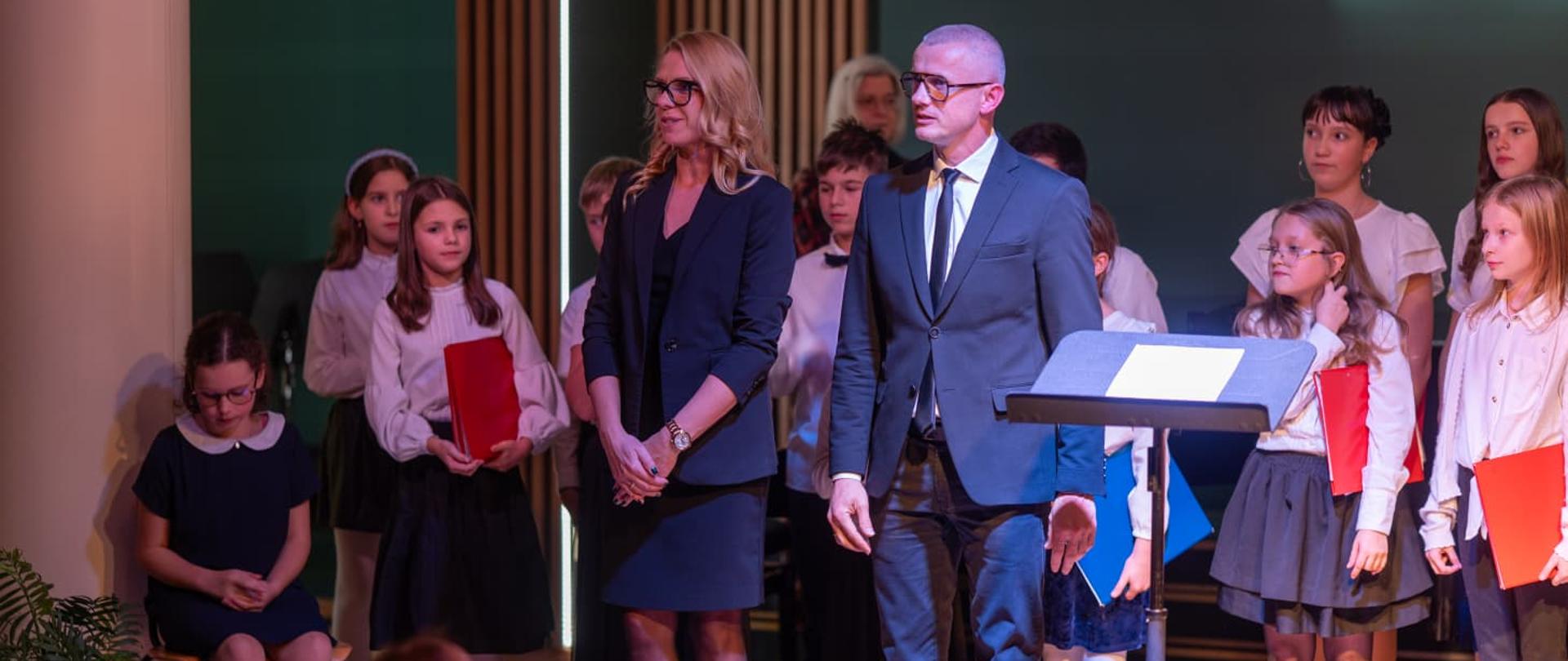 Na zdjęciu i widoczne są dzieci stojące na scenie w sali koncertowej są ubrane w stroje galowe a w rękach trzymają czerwone teczki. W tle widoczna jest ściana w kolorze zielonym. Przed dziećmi widać 2 prowadzących koncert