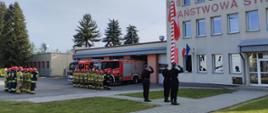Na placu przed Jednostka Ratowniczo Gaśnicza nr 1 stoją strażacy w dwóch szpalerach naprzeciw siebie na uroczystej zbiórce. Pomiędzy szpalerami stoi dowódca. Strażacy ubrani są w stroje bojowe. Za drugim szpalerem widoczne są samochody gaśnicze. Na maszt uroczyście wciągana jest biało czerwona flaga.