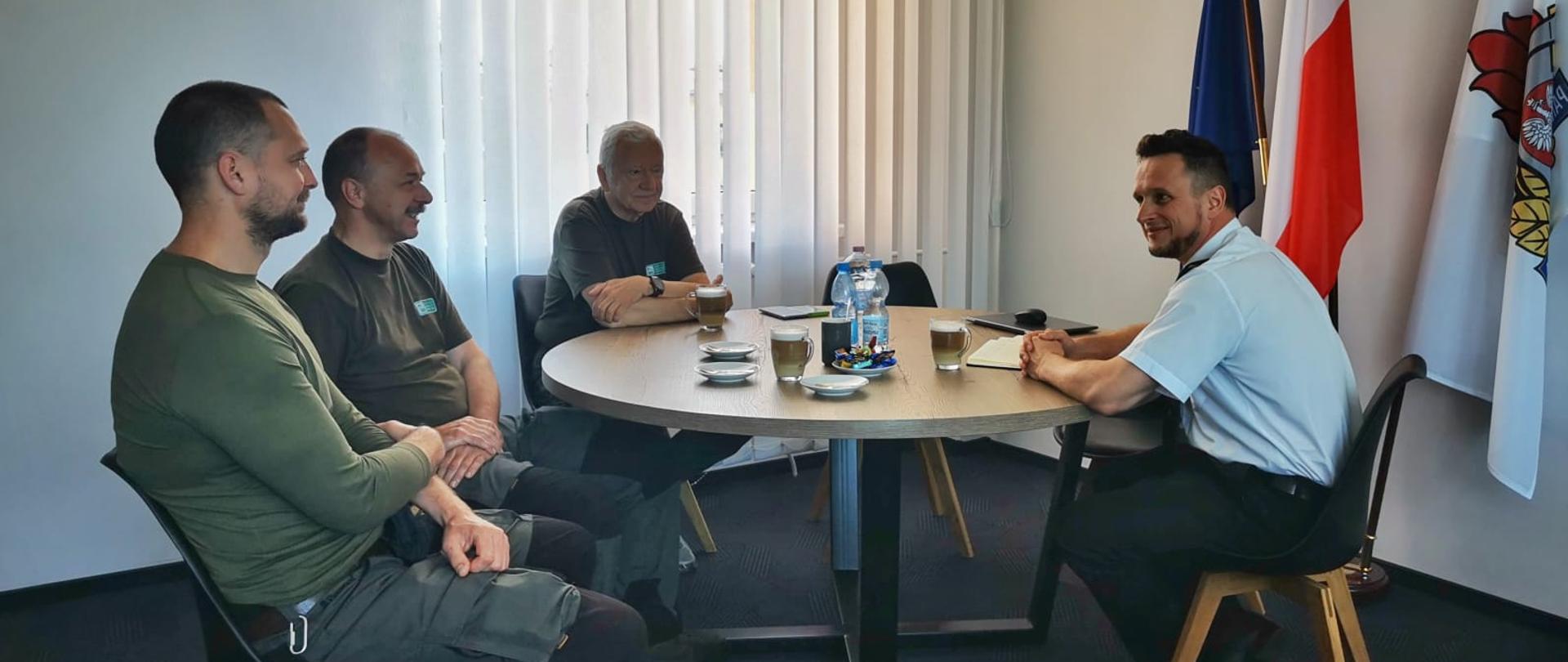 W gabinecie przy stole siedzą Komendant Powiatowy PSP w Hajnówce bryg. Koszczuk Piotr i członkowie zespołu w zielonych mundurach.