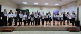 Uczniowie klas pierwszych cyklu czteroletniego Państwowej Szkoły Muzycznej I stopnia im. Jana Sebastiana Bacha w Grajewie stoją na scenie z dyplomami, obok nauczycieli. Uczestnicy ubrani są w stroje galowe(białe koszule i czarne spódnice/spodnie). W tle widoczne są fortepian, godło Polski oraz baner szkoły