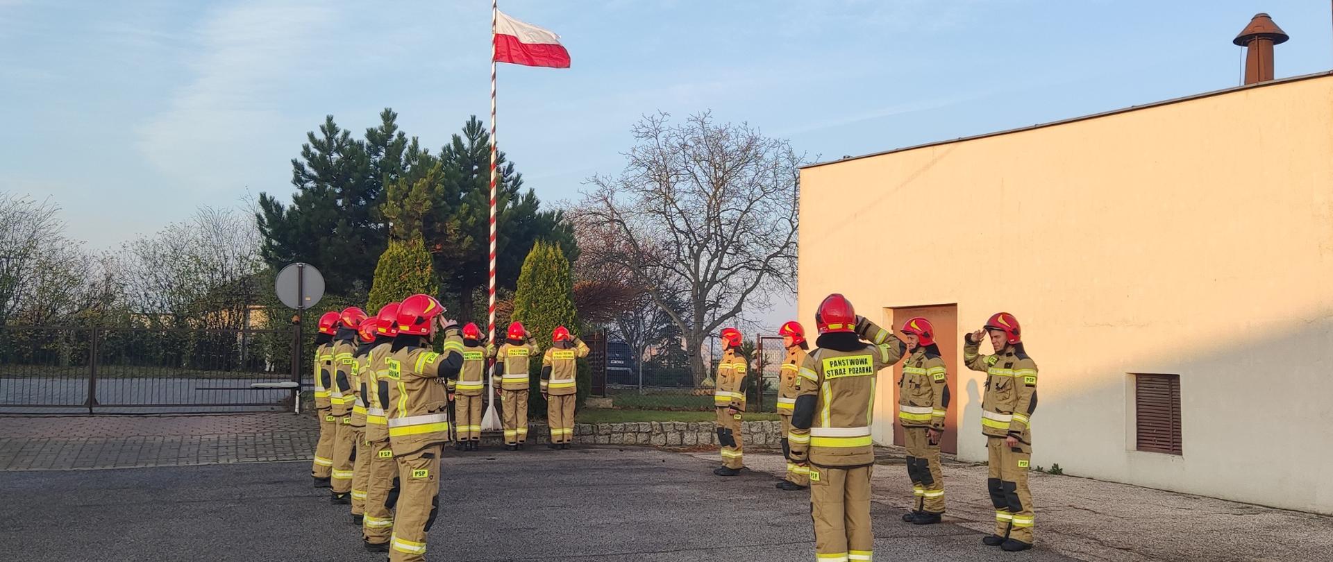 Zdjęcie przedstawia: Plac Komendy Powiatowej Państwowej Straży Pożarnej na którym odbywa się uroczysta zbiórka. Strażacy ubrani w ubabrania specjalne koloru piaskowego oraz czerwone hełmy zawieszają flagę Państwową i odśpiewują hymn narodowy.