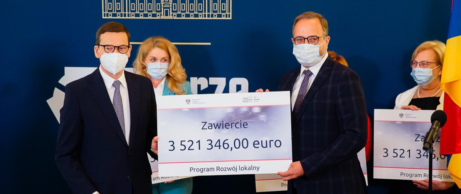 Premier Mateusz Morawiecki stoi, obok włodarze miast trzymają w rękach czeki.