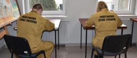 Zdjęcie przedstawia dwóch strażaków siedzących tyłem do fotografa przy stoliku w żółtych mundurach z napisem Państwowa Straż Pożarna