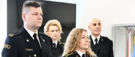 Dwóch strażaków w mundurach wyjściowych stoi obok siebie mężczyzna kobieta za nimi stoi dwóch funkcjonariuszy w mundurach wyjściowych za nimi ustawiony jest ekran oraz sztandar w gablocie.